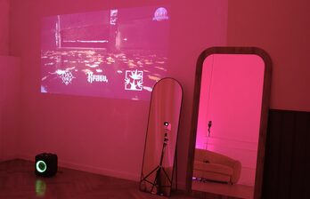 Комната с проектором на стене, показывающим красочное изображение. На полу черная колонка. Два больших зеркала, одно с деревянной рамой, другое на треноге. Слабое розовое освещение, диван виден в отражении зеркала. Паркетный пол.