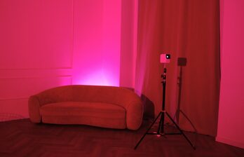 Пространство с розовой подсветкой, диван плавных форм, штатив с маленьким проектором, шторы. Пол из темного дерева, стена светлая с декоративными элементами.