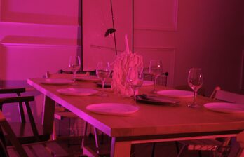 Столовая в розовом освещении, деревянный стол с шестью накрытыми приборами, бокалы для вина, декоративная композиция по центру. Стулья разных дизайнов вокруг стола. Белые наклонные панели на стенах.