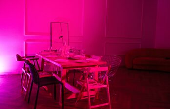 Просторная комната с деревянным полом, розовым освещением. В центре деревянный стол с посудой и бокалами, окруженный разноцветными стульями. Слева зеркало, справа коричневый диван. Стены украшены панельным декором.