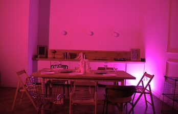 Столовая зона с деревянным столом, окруженным разными стульями, на столе тарелки и бокалы. На заднем плане открытые полки с предметами и декоративное освещение розового цвета. Белые стены, паркетный пол. Трековые светильники на стене.