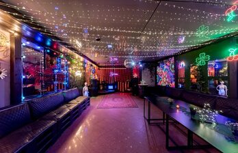 Просторное помещение с длинными диванами вдоль стен, низкими столами и цветными неоновыми украшениями на стенах. Потолок украшен множеством маленьких светодиодов. Пол гладкий, с розовым ковром. Зеркала на стенах, приглушенное освещение.