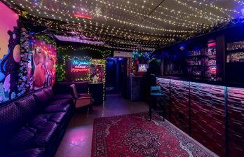 Лаунж-пространство с барной стойкой, черной кожаной мебелью и деревянными элементами отделки. Потолок украшен гирляндами. На стене яркие граффити и неоновая вывеска. Ковровое покрытие с орнаментом на полу.
