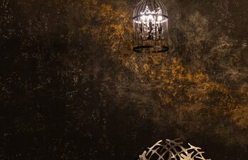 Темная стена с текстурой и золотистыми акцентами, подвесной светильник в форме клетки с лампой внутри, металлический декоративный элемент на переднем плане. Освещение теплое и направленное.