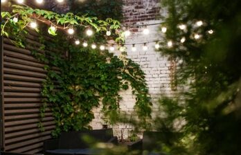 Открытое пространство с кирпичными стенами, покрытыми растениями. Струнные лампочки натянуты над зоной. Плетеная мебель темных цветов. Один угол имеет деревянную решётку. Освещение теплое, атмосфера уютная.