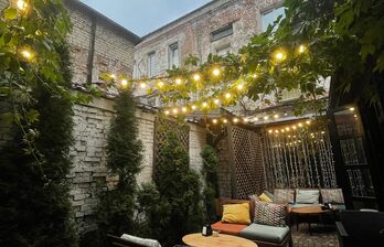 Уличное пространство с кирпичными стенами, выложенной плиткой площадкой и диванами с подушками. Множество растений и гирлянд с лампочками. Деревянные круглые столы, мягкие сиденья, разноцветные подушки. Вертикальные решетки с растениями.