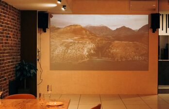 Помещение с кирпичной стеной слева и гладкой стеной, на которой проецируется изображение горного пейзажа. Пол с крупной плиткой. Стол из дерева с двумя бокалами и пустой тарелкой. Потолочные светильники обеспечивают мягкое освещение.