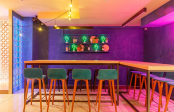 Барное пространство с высоким столом и стульями с зелеными сиденьями. Фиолетовая стена с полкой, на которой расположены неоновые фигуры кактусов и бутылки. Яркое освещение с теплым и фиолетовым оттенком, плиточный пол.