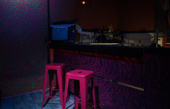 Темное помещение с барной стойкой, за которой стоят два розовых стула. На стойке расположены бокалы и старый телевизор с кружевной салфеткой. Стены обшиты темными обоями с узором. Освещение слабо-оранжевое, пол темного дерева.