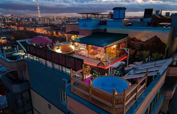 Крыша здания с террасой, огороженной перилами. На террасе находится круглая деревянная купель с водой, столы и стулья под зонтом. Присутствует декоративное освещение, стеклянная перегородка, стена с изображением гор. Вдали виден городской пейзаж.