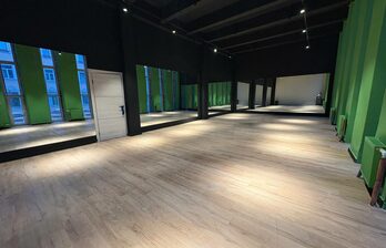 Просторное помещение с деревянным полом, стены в зелёных и тёмных оттенках, большие окна с отражением домов, светодиодное освещение на потолке, белая дверь сбоку.