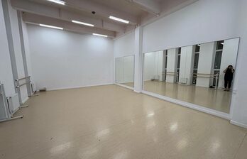 Просторный зал с бежевым полом, белыми стенами и высокими потолками. Большое зеркало на одной из стен. Установлены светильники на потолке. Справа видны окна и несколько хореографических станков.