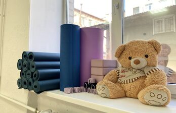 Подоконник с йога-ковриками синего и фиолетового цвета, мягкими блоками и ремнями для йоги, плюшевый медведь в полосатой кофте. Белая рама окна, вид на городские здания. Светлый интерьер.