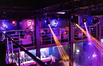 Двухуровневое пространство с балконом, оборудованное барной стойкой. Яркое цветное освещение, пара кресел у стен с картинами. Металлические перила и конструкции. Люди за баром. Световые лучи отражаются от поверхностей.