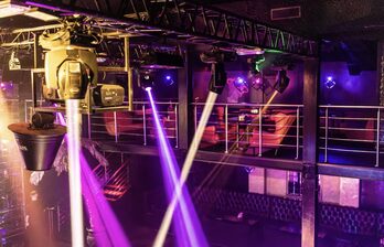 Клубное пространство с балконом, оборудованное софитами и прожекторами. Диваны красного цвета вдоль стен, металлические перила на втором уровне. Многоцветное освещение, включая фиолетовые и розовые лучи. Потолочные конструкции и сцена.