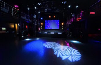 Большое тёмное помещение с сценой и экраном. Металлические балконы по бокам, диско-шары, разноцветное освещение с узорами на полу. Черный пол, высокий потолок, световые приборы, цветная подсветка. Монохромная и фиолетовая гамма.
