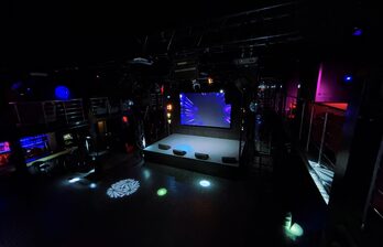 Ночной клуб с двумя уровнями и сценой. Центральный экран, несколько световых эффектов на полу. Оборудование для света и звука. Барная стойка слева, металлические перила вдоль второго уровня. Черный интерьер с разноцветной светодиодной подсветкой.
