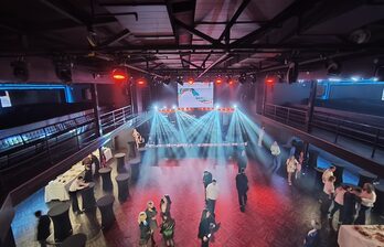 Просторная комната с высокими потолками, оснащена сценой с подсветкой и большим экраном. Вокруг расставлены высокие круглые столы с белыми скатертями. На полу паркет, по периметру полумрак с ярким светом на сцене.