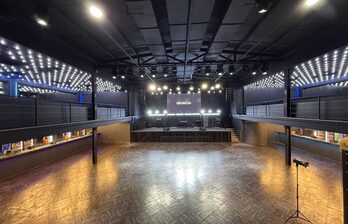 Большое открытое пространство с паркетным полом. Высокий потолок с черными балками и подвесными светильниками. Центральная сцена с экраном и музыкальным оборудованием. По периметру балконы с поручнями. Интенсивное потолочное освещение.