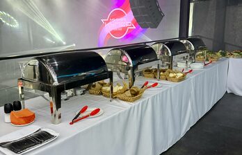 Помещение для банкета с длинным столом, покрытым белой скатертью. На столе четыре металлических контейнера для еды, подносы с хлебом, красные щипцы, посуда, посолонки и салфетки. На заднем плане светодиодные панели с текстом.