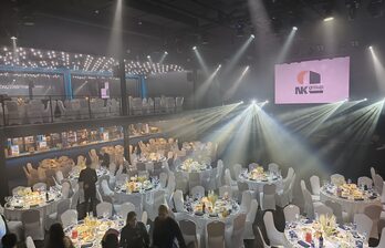 Большой зал с круглым столами, покрытыми скатертями; на каждом столе свечи и сервировка. Белые стулья, световые лучи по залу. Двухуровневое пространство, на верхнем уровне также столы и стулья. На стене висит экран с логотипом.