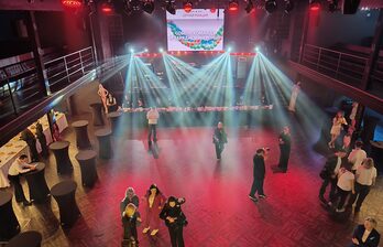 Большое внутреннее пространство с деревянным полом и балконами на втором уровне. На переднем плане высокие круглые столы. На заднем плане сцена с экраном и световыми эффектами. Люди стоят и общаются. Освещение разноцветное.