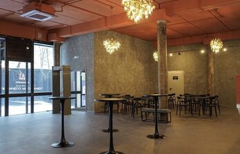 Просторное помещение с выполненными в индустриальном стиле стенами, равномерно расположенными столиками и стульями. Выделяются стеклянные двери, декоративные светильники и ярко оранжевый потолок с видимой вентиляцией.