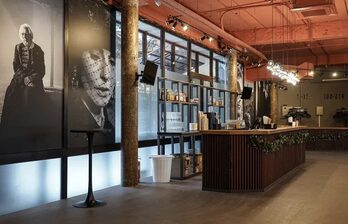 Барное пространство в индустриальном стиле с бетонными колоннами и деревянной стойкой. Большие чёрно-белые портреты украшают стены. На потолке открытые коммуникации. Освещение представлено подвесными лампами. Полки с бутылками, мусорная корзина.