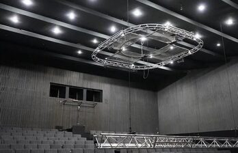 Зал с амфитеатром, серые ступенчатые ряды кресел, тёмный рельефный потолок с лампами, подвесная металлическая конструкция, стены отделаны вертикальными панелями серого цвета.