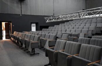 Аудиторный зал с рядной расстановкой серых кресел с подлокотниками. Пол покрыт ковролином, стены темные. Над креслами металлическая конструкция для освещения. Справа от двери виден шкаф. Освещение из дверного проема.