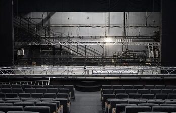 Аудитория со сценой и рядами темных кресел. На сцене установлена металлическая конструкция. Задняя стена с серыми панелями и трубами. Освещение тусклое, фон серый.