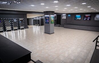 Просторное помещение с плиточным полом и серыми стенами. На стенах размещены картины. Вдоль стены стоят зеркала с подсветкой. Центральная колонна украшена изображениями. Потолок оснащен встроенными светильниками.
