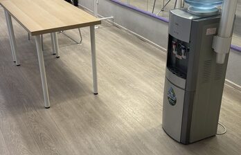 Пространство с деревянным столом и кулером для воды. Светлое напольное покрытие, серые стены, зеркало вдоль стены. Освещение естественное, мебель минималистичная. Электрическая розетка и провода на стене.