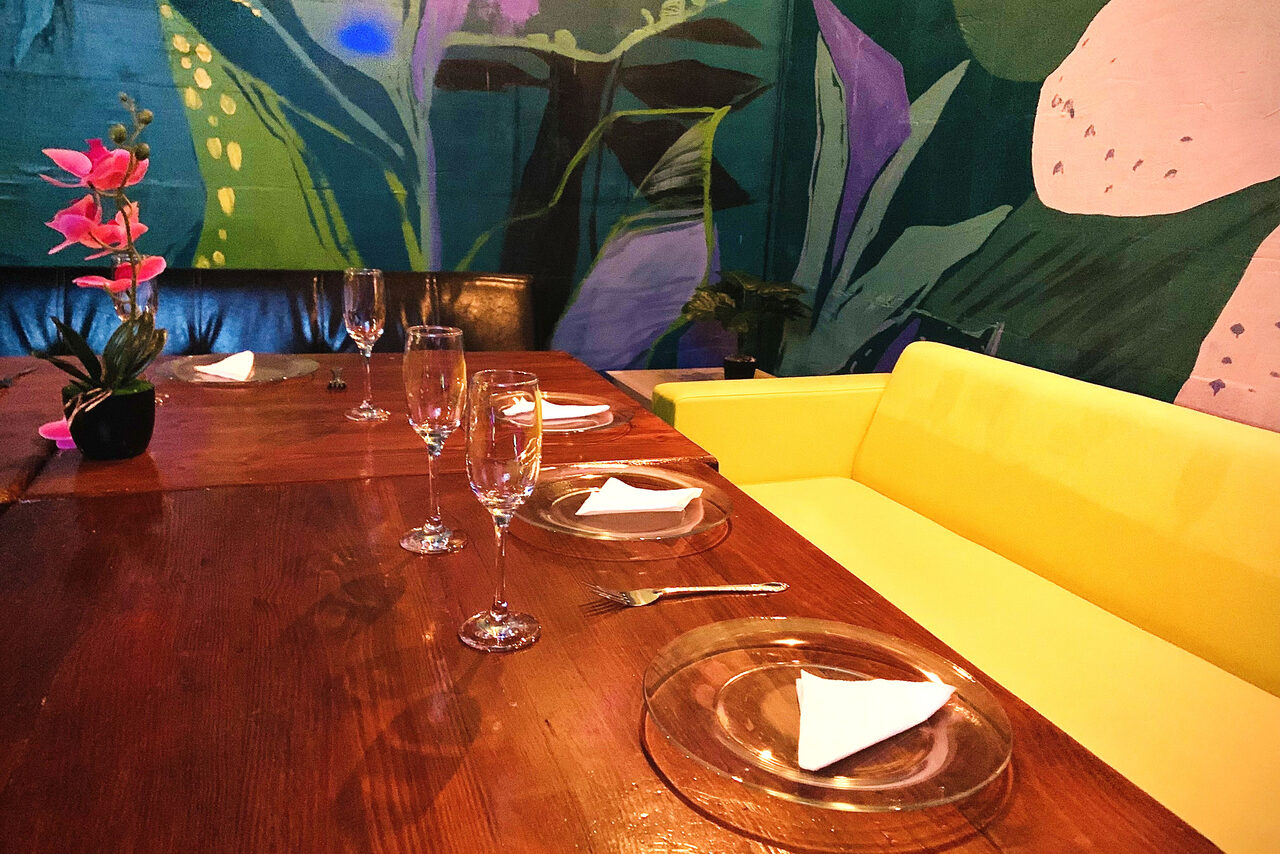 Арендовать Уютный тропический лофт. Бокалы и тарелки с вилками на столе. Небольшой цветок в горшке. Разноцветные диваны
