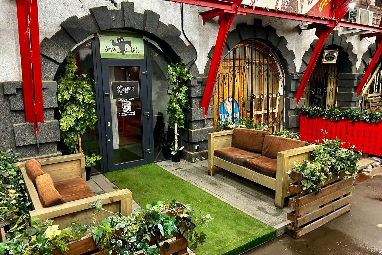Арендовать Creative space with a bar counter. Вход в пространство со стеклянной дверью; арка декорирована зеленью. В зоне перед входом две деревянные диваны с коричневыми подушками. Искусственная трава на полу, деревянные кашпо с растениями. Серо-красная отделка стен.