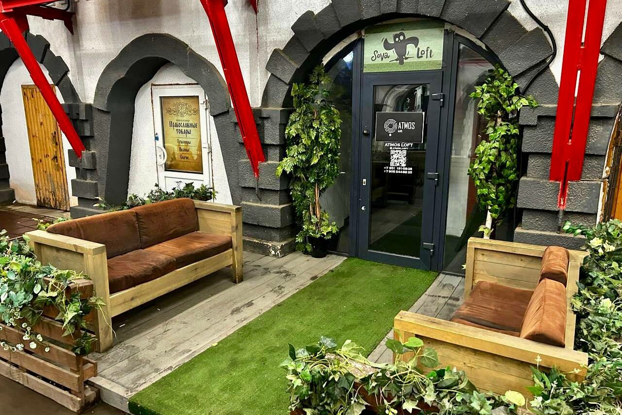Арендовать Creative space with a bar counter. Вход в помещение с каменными арками, двумя деревянными диванами с коричневыми подушками, искусственной зелёной травой и растениями. Дверь стеклянная с наклейками. Стены белые с серыми элементами и красными металлическими балками.
