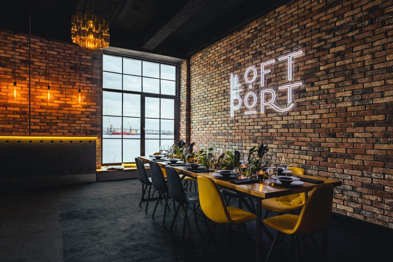 Арендовать Лофт Порт. Loft-пространство с кирпичными стенами, длинным обеденным столом и разноцветными стульями. На стене световая надпись. Большое окно с видом на воду. Подвесные светильники и золотистая люстра. Стены обшиты деревом, пол темный.