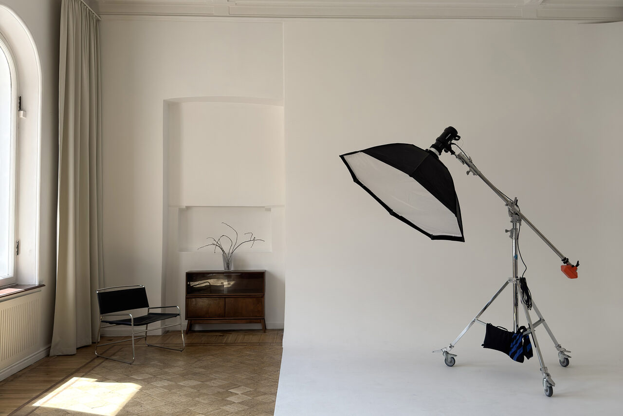 Арендовать Bedford studio (Бедфорд Студия). Светлый зал с белой циклорамой и балконом. Фотостудия с белыми стенами и светлым паркетным полом. На переднем плане софтбокс на стойке с колесами. Справа кресло, слева тумба с растением. Большое окно с занавесями. Пространство минималистично и светло.