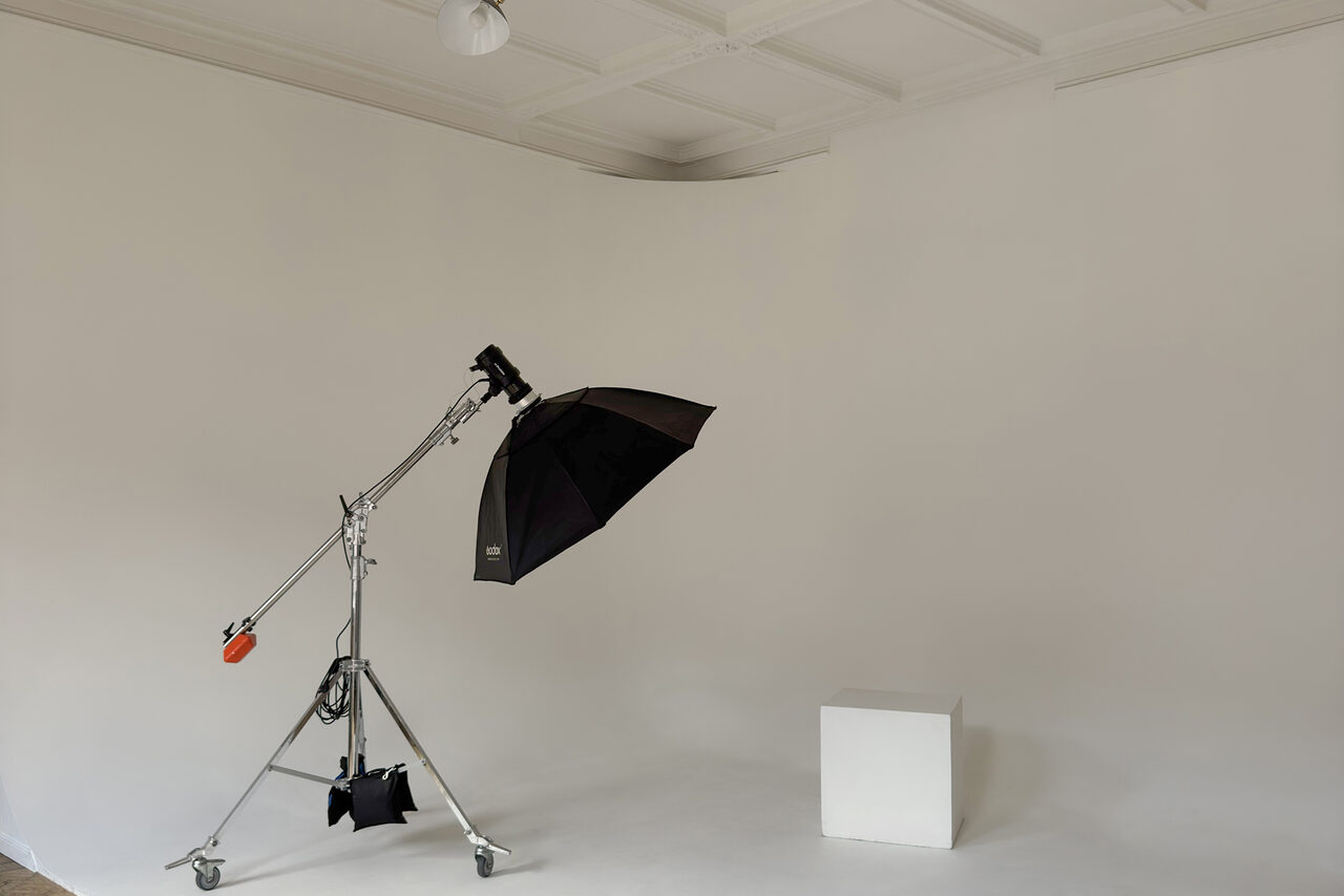 Арендовать Bedford studio (Бедфорд Студия). Светлый зал с белой циклорамой и балконом. Фотостудия с белыми стенами и белым полом. Светло-серое мягкое освещение. В центре студийный осветитель с черным зонтом на стойке. Справа белый куб. Потолок с квадратной панельной отделкой.