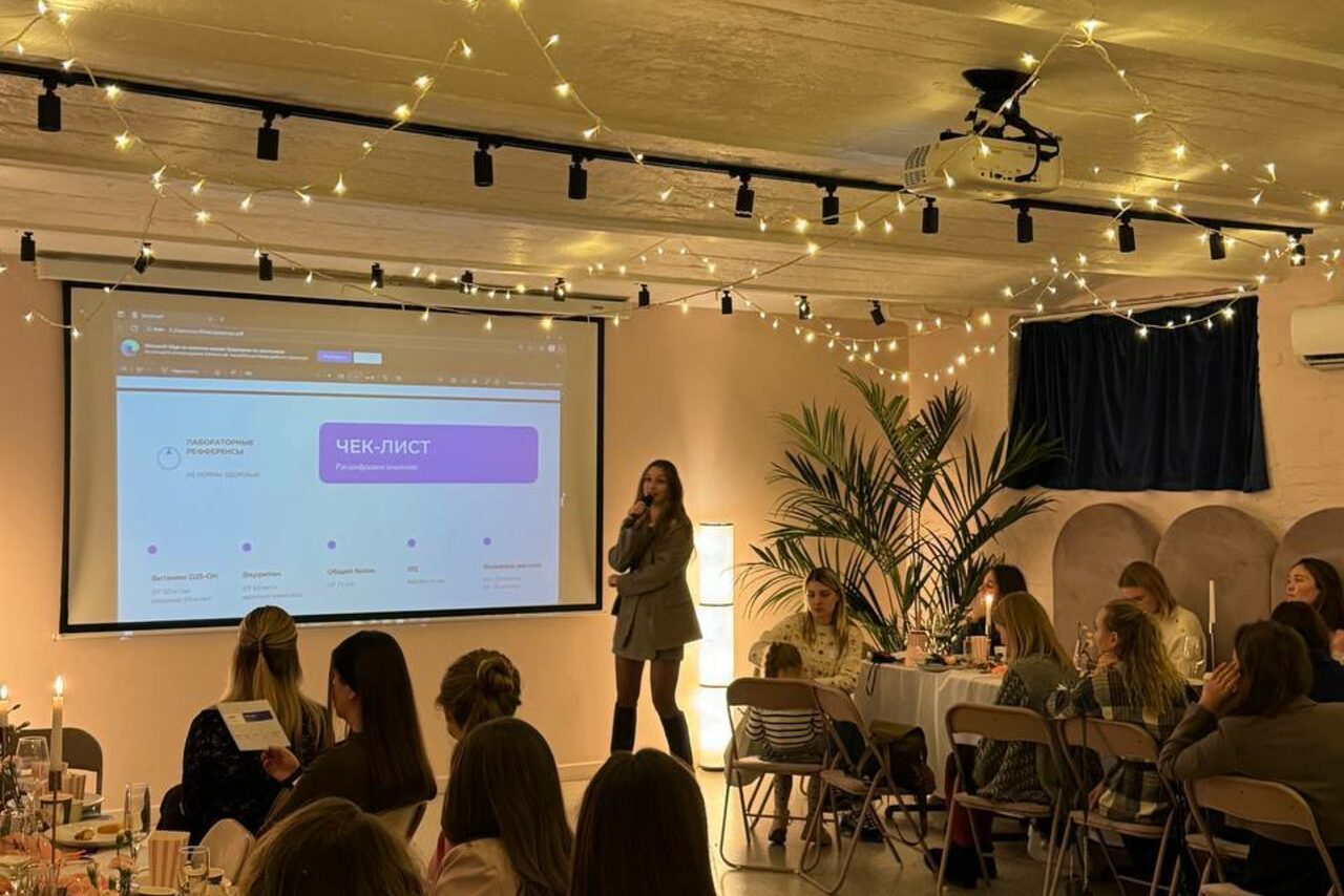 Арендовать Стилизованный лофт в сине-розовых оттенках. Просторная комната с низким потолком украшена гирляндами и занавеской. Установлен проектор с экраном, на экране текст "Чек-лист". Спикер выступает перед аудиторией, сидящей за светлыми столами на складных стульях. В углу крупное растение.