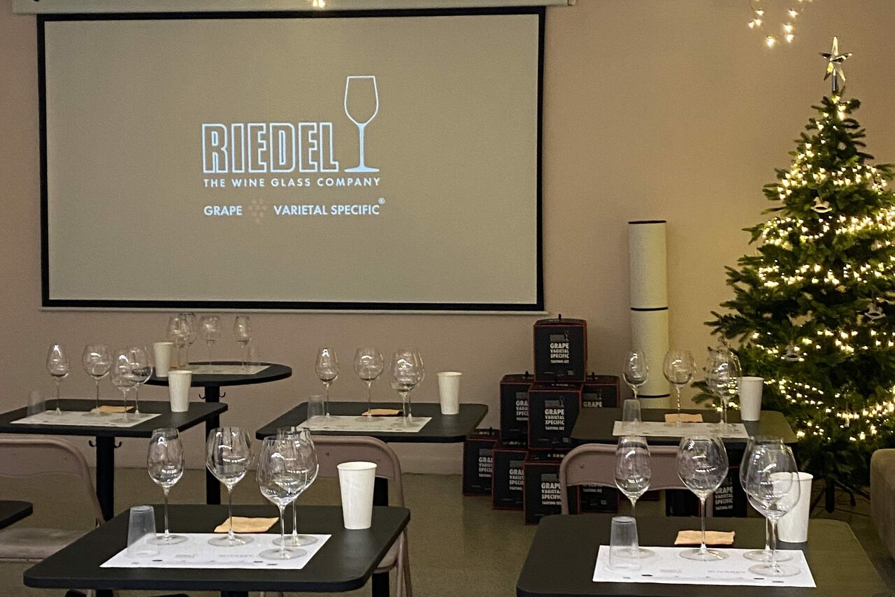 Арендовать Стилизованный лофт в сине-розовых оттенках. Просторная комната с проектором на стене. Ряды столов с бокалами, стаканами и бумажными стаканчиками. Справа украшенная елка с гирляндой. Спокойные тона интерьера. На экране текст: "Riedel The Wine Glass Company Grape Varietal Specific".