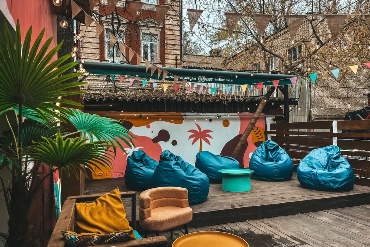 Арендовать Лофт Surf (Серф). Тропический лофт с большой верандой.. Уличная площадка с деревянным настилом, синими креслами-мешками, круглым бирюзовым столом, бежевым креслом и желтыми подушками. Разноцветные флажки над головой, стена с ярким граффити, зелёные растения.
