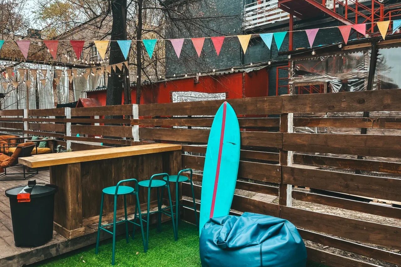 Арендовать Лофт Surf (Серф). Тропический лофт с большой верандой.. Открытая площадка с деревянным забором и барной стойкой. Три высоких стула, синий пуф и вертикально стоящая доска. Искусственная трава на земле. Над пространством висят гирлянды из разноцветных флажков. На заднем плане видны здания.