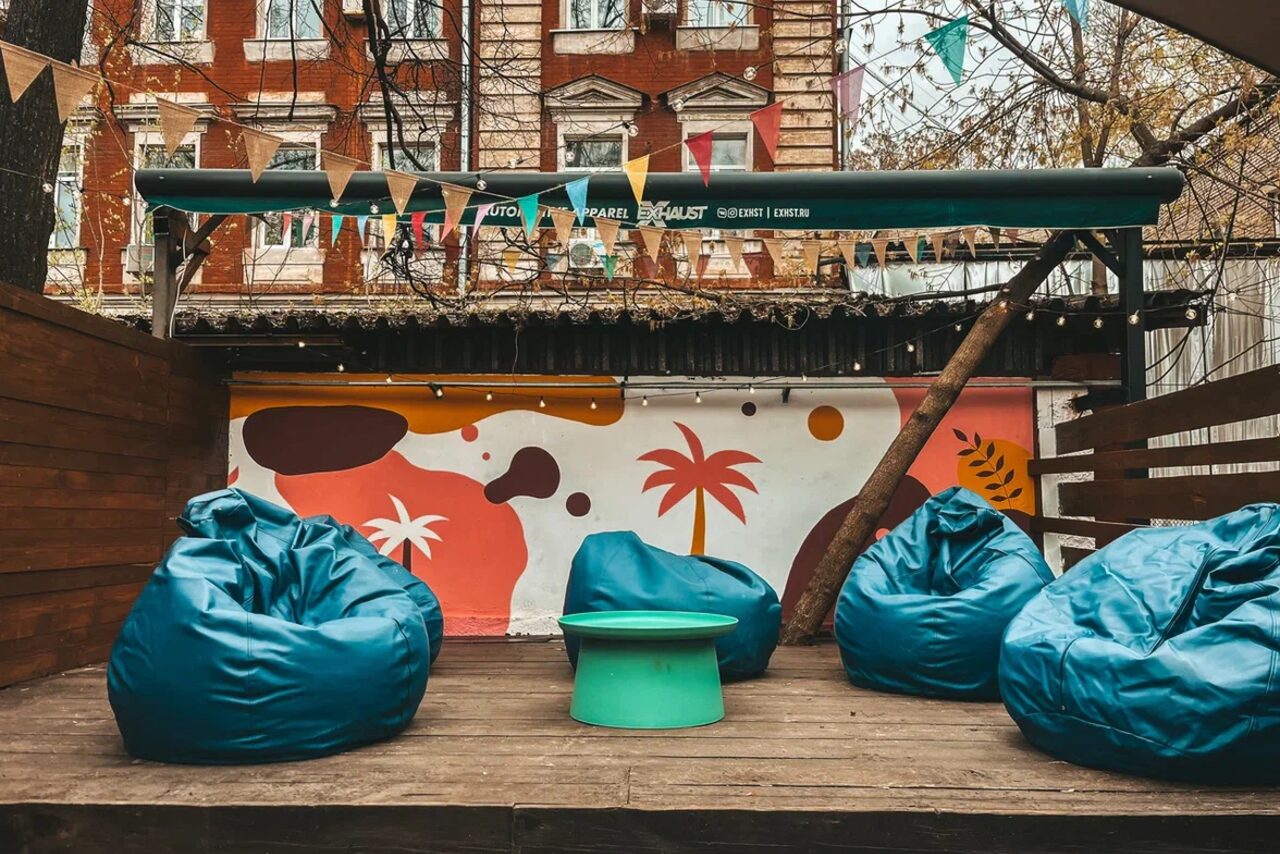 Арендовать Лофт Surf (Серф). Тропический лофт с большой верандой.. Открытая терраса с деревянным полом, четыре синих кресла-мешка, зеленый круглый столик, декоративная цветная стена с рисунками пальм, деревянные перегородки, подвешены разноцветные флажки, надписи на навесе.