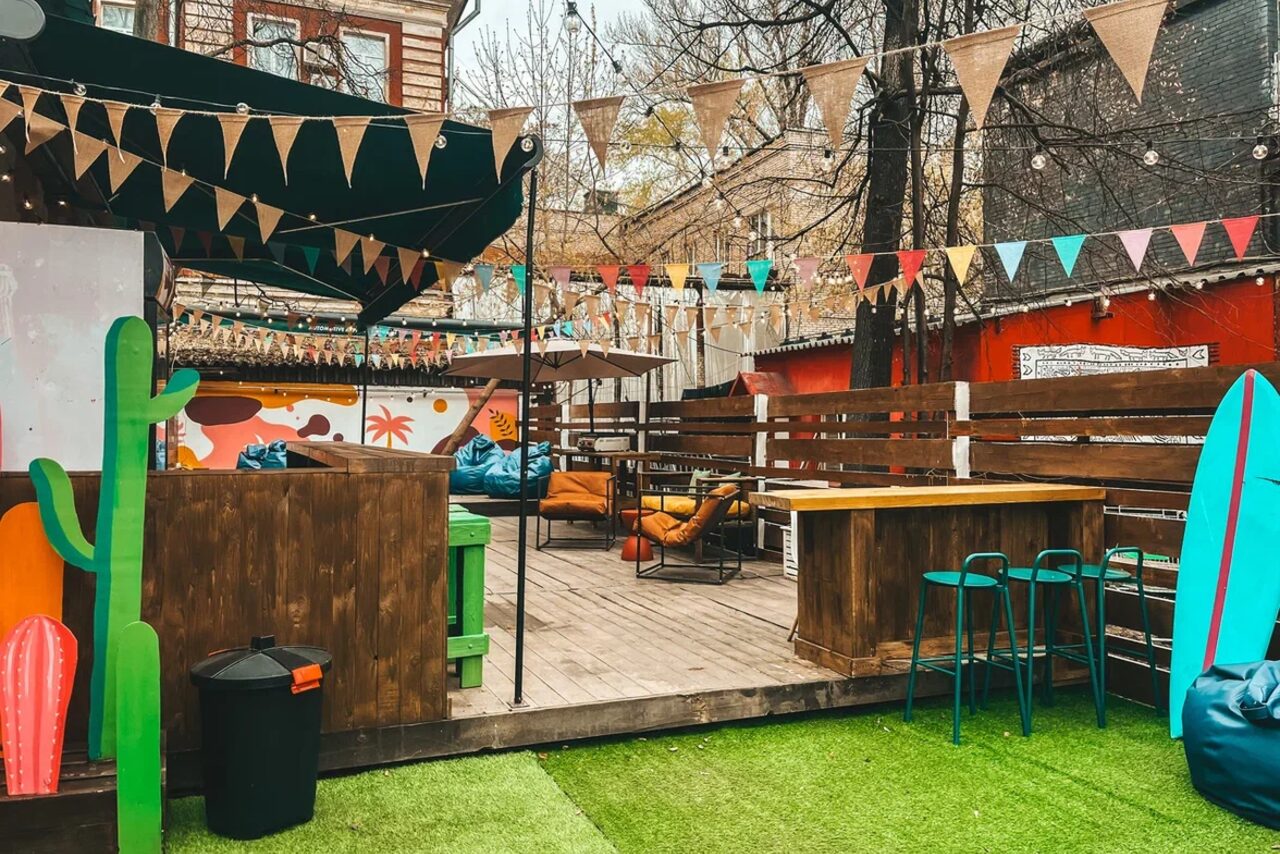 Арендовать Лофт Surf (Серф). Тропический лофт с большой верандой.. Открытая терраса с деревянным настилом, барной стойкой и высокими стульями. Разноцветные флажки и гирлянды. Зеленый искусственный газон. Яркие диваны и подушки. На стене граффити. Праздничное оформление.