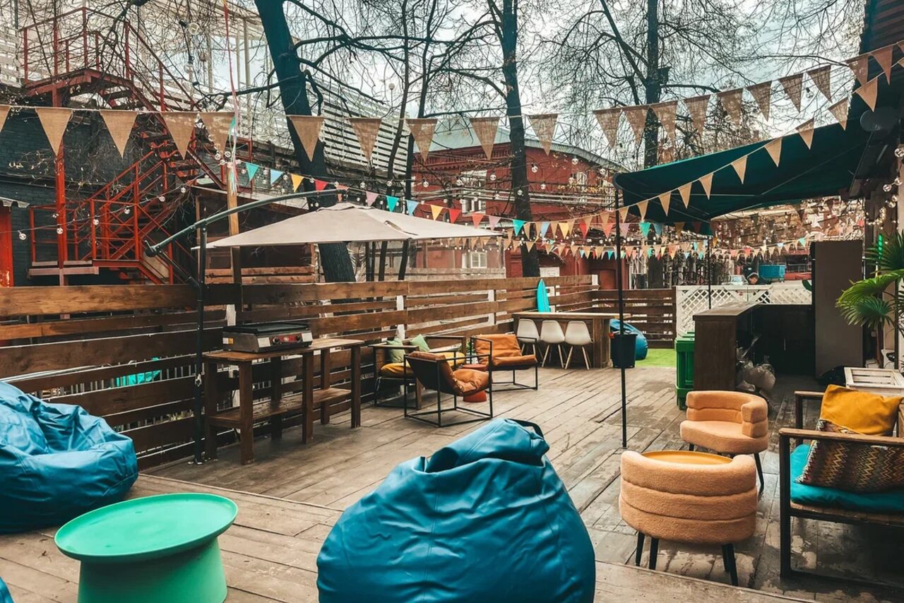 Арендовать Лофт Surf (Серф). Тропический лофт с большой верандой.. Открытая терраса с деревянным настилом, столами и стульями. Несколько кресел и синие мешки для сидения. Над пространством висят разноцветные флажки. На заднем плане деревья и лестница.