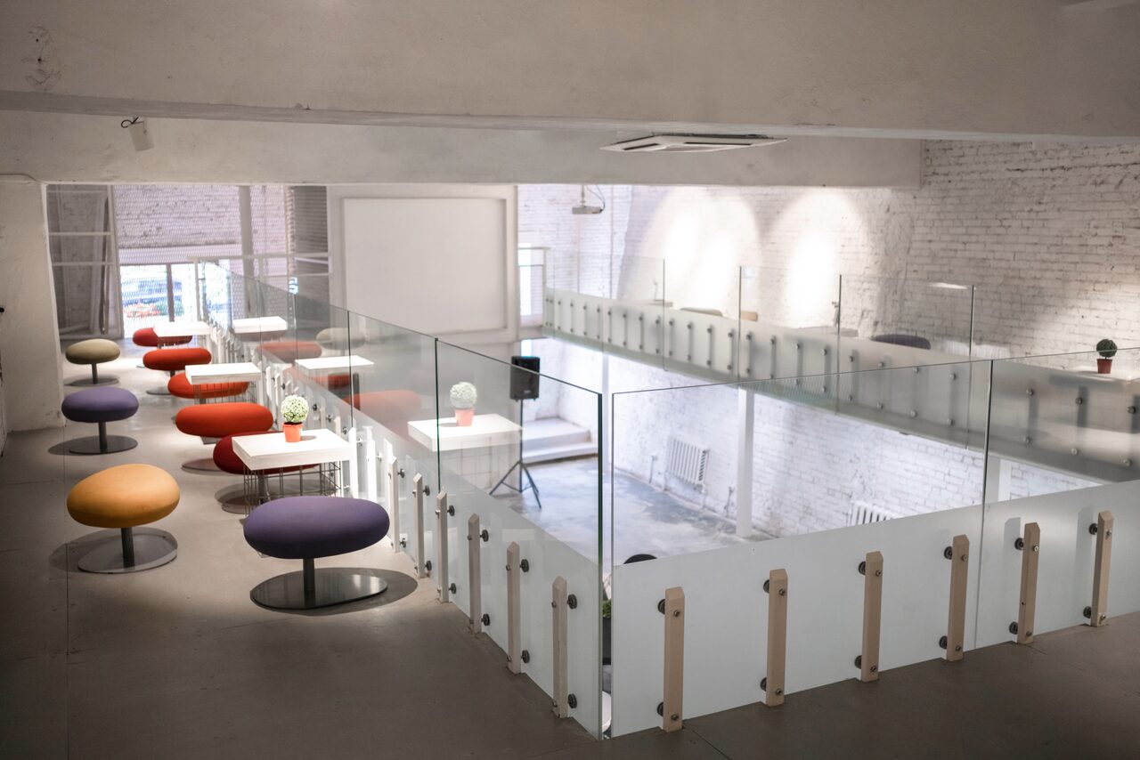Арендовать AG Loft (АГ Лофт). Белый лофт с галереей-балконом и верандой. Простор с белыми кирпичными стенами и серым полом. Мезонин со стальной и стеклянной оградой. Разноцветные круглые пуфы и круглые белые столы. Яркое потолочное освещение. Зона с белым экраном. Большие окна с естественным светом.
