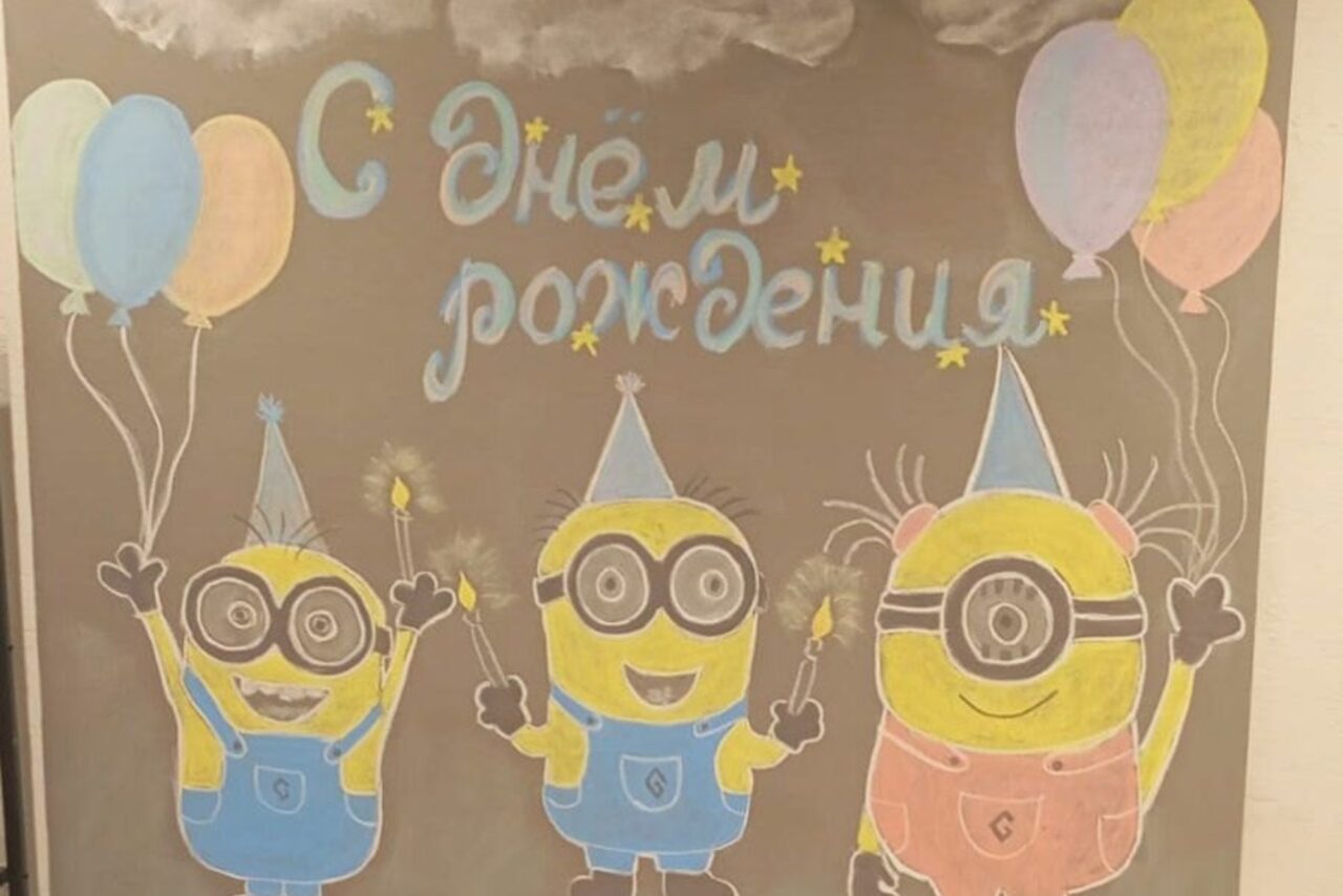 Арендовать Уютный лофт с меловой стеной. Чёрная доска с нарисованными мелом персонажами в синих и жёлтых цветах. Надпись "С днём рождения" сверху, окружена воздушными шарами. Персонажи нарисованы в праздничных колпаках с бенгальскими огнями в руках.