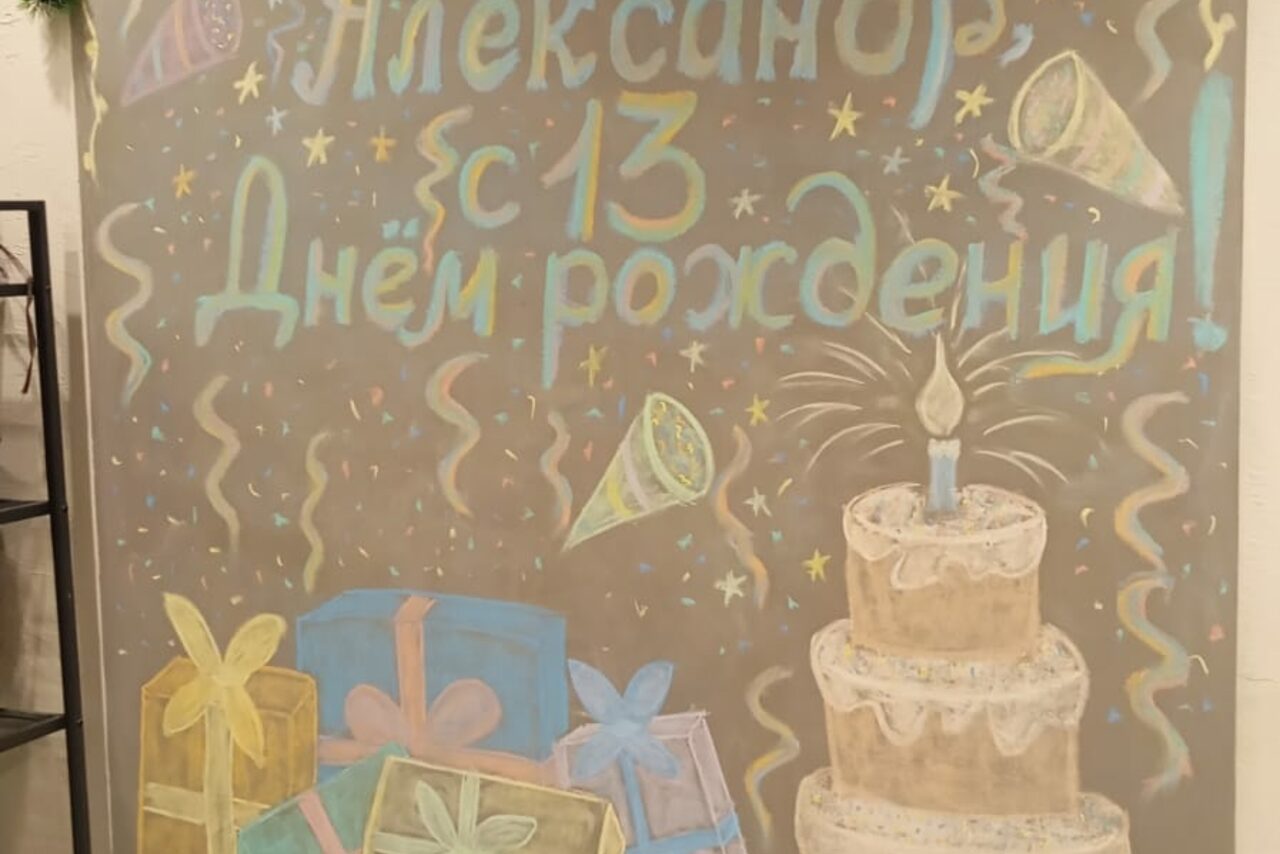 Арендовать Уютный лофт с меловой стеной. Настенная роспись с поздравительной надписью и изображениями свечи, торта, подарков и серпантинов на сером фоне. Текст большой, разноцветный. Оттенки: синий, жёлтый, розовый. Атмосфера праздничная.