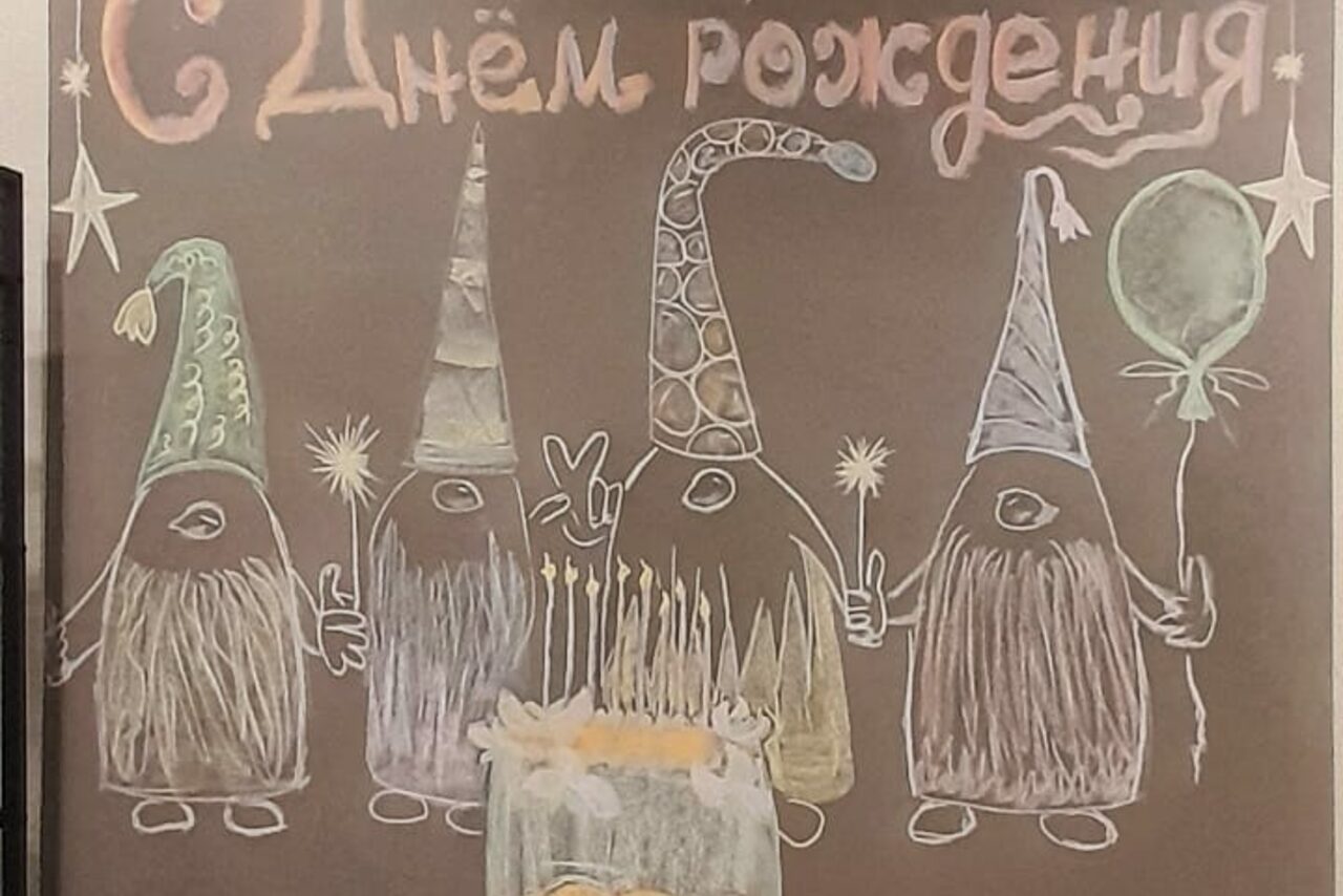 Арендовать Уютный лофт с меловой стеной. Доска с мелками, на которой нарисованы четыре гнома в колпаках, держащие свечи и воздушный шар. Перед ними праздничный торт со свечами. Вверху надпись "С Днем рождения". Фон коричневый, рисунки разноцветные.